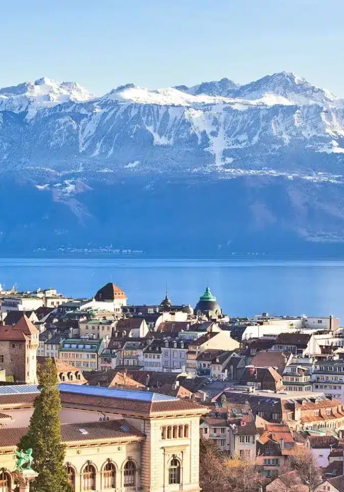Vue panoramique de Lausanne, en Suisse, montrant des bâtiments historiques, une cathédrale, le lac Léman et les Alpes enneigées en arrière-plan, sous un ciel bleu clair.