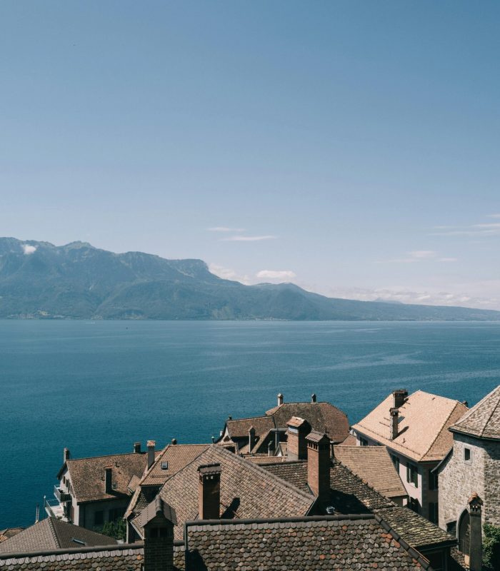 Les toits des maisons en pierre surplombent un lac bleu calme avec des montagnes au loin sous un ciel clair.