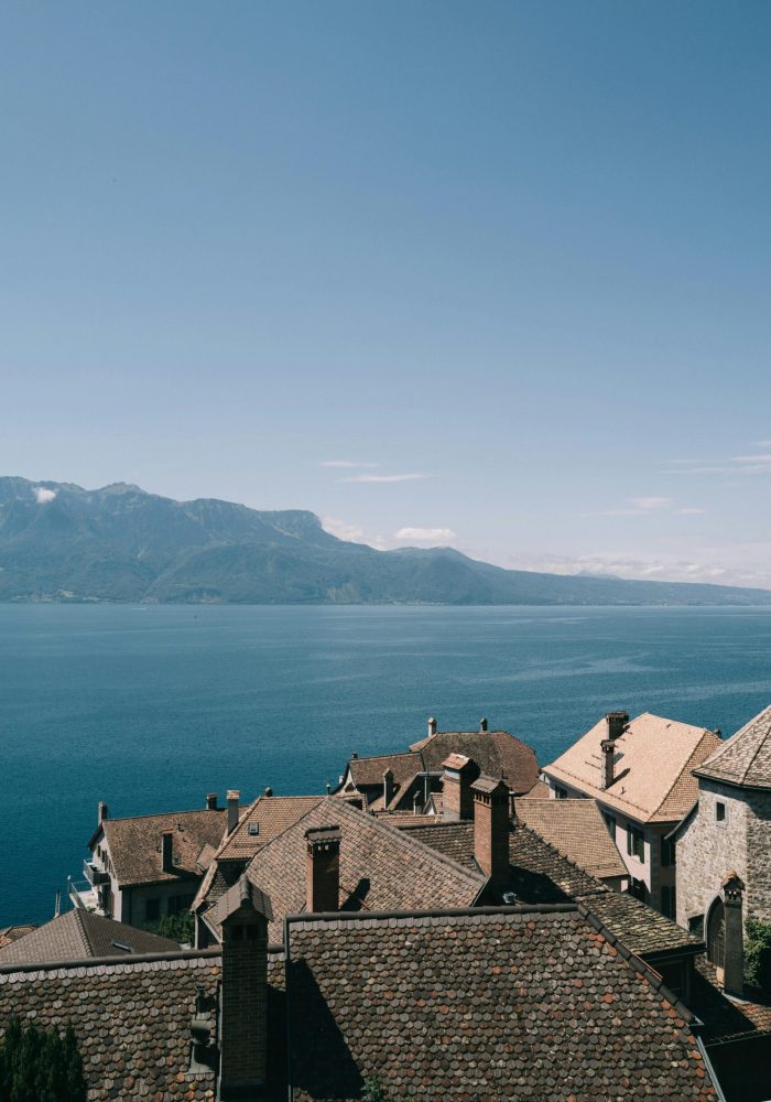 Les toits des maisons en pierre surplombent un lac bleu calme avec des montagnes au loin sous un ciel clair.