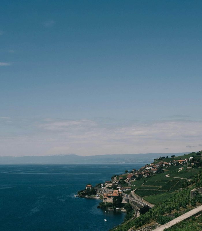 Un village côtier avec des maisons et des vignobles s'étend le long d'un rivage incurvé, au bord d'une eau d'un bleu profond, sous un ciel clair et ensoleillé. Une route sinueuse est parallèle à la côte, avec des collines en arrière-plan.