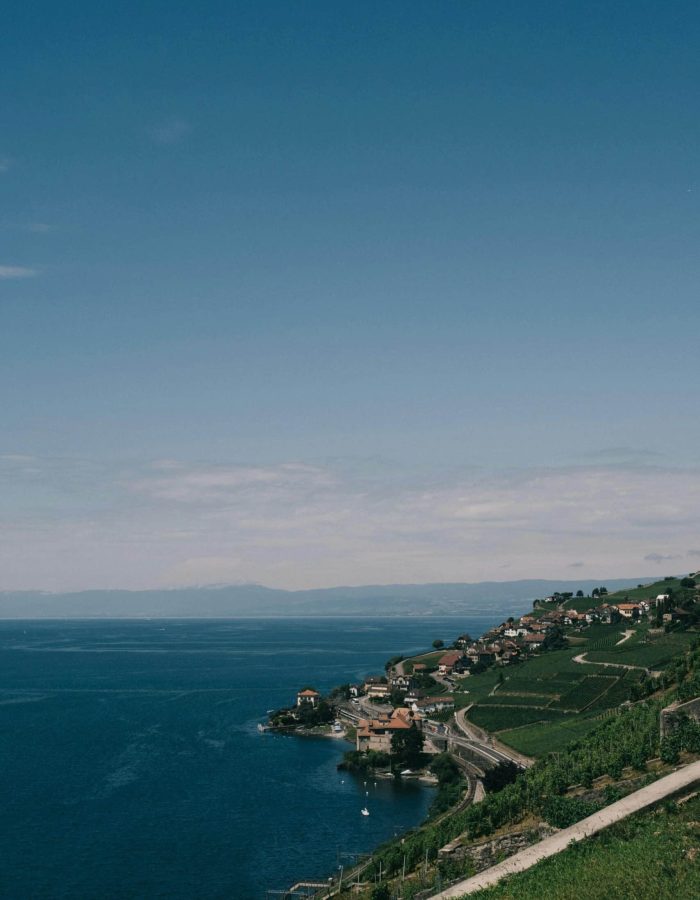 Un village côtier avec des maisons et des vignobles s'étend le long d'un rivage incurvé, au bord d'une eau d'un bleu profond, sous un ciel clair et ensoleillé. Une route sinueuse est parallèle à la côte, avec des collines en arrière-plan.