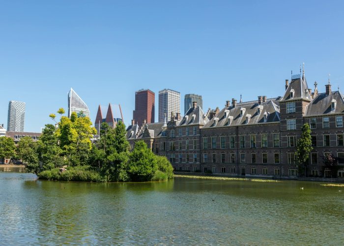 Un bâtiment historique aux toits pointus se dresse à côté d'un étang calme avec une petite île couverte d'arbres, tandis que des gratte-ciel modernes s'élèvent à l'arrière-plan sous un ciel bleu limpide.