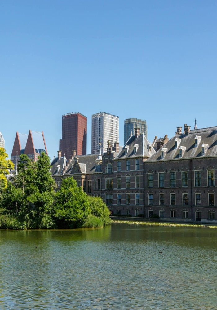 Un bâtiment historique aux toits pointus se dresse à côté d'un étang calme avec une petite île couverte d'arbres, tandis que des gratte-ciel modernes s'élèvent à l'arrière-plan sous un ciel bleu limpide.