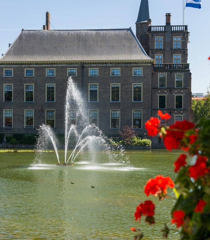 Un grand bâtiment en briques avec de nombreuses fenêtres se dresse derrière un étang avec une fontaine pulvérisant de l'eau. Des fleurs rouges et des feuilles vertes se trouvent au premier plan. Un drapeau bleu flotte sur le bâtiment sous un ciel bleu clair.