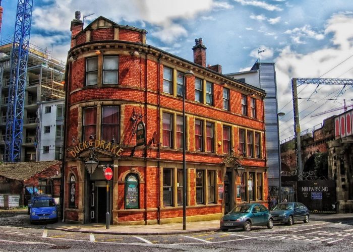 Un pub historique en briques rouges appelé "Duck & Drake" se trouve au coin d'une rue, avec des fenêtres cintrées, deux cheminées et plusieurs voitures garées à proximité. Des bâtiments modernes et un pont ferroviaire sont visibles à l'arrière-plan.