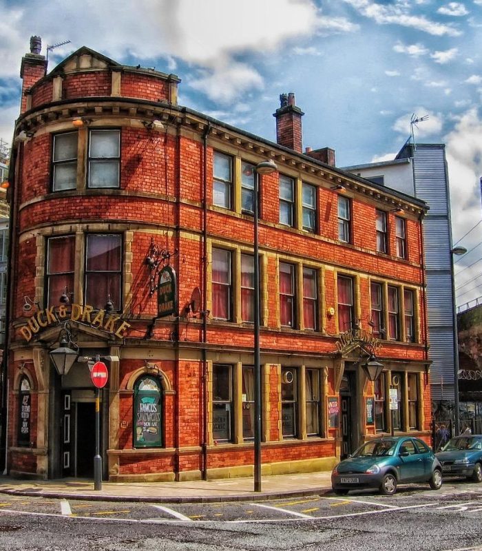 A historic red brick pub called "Duck & Drake" stands on a street corner, with arched windows, two fireplaces and several cars parked nearby. Modern buildings and a railway bridge can be seen in the background.