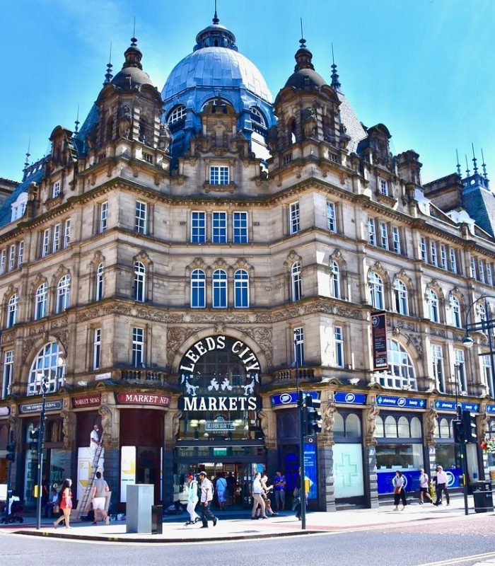 Un grand bâtiment historique orné d'un toit en dôme abrite les marchés de la ville de Leeds. Les gens marchent le long du trottoir et les boutiques s'alignent au rez-de-chaussée sous un ciel bleu éclatant.
