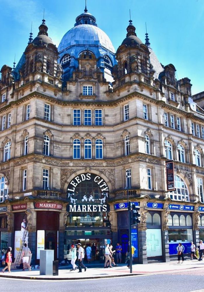 Un grand bâtiment historique orné d'un toit en dôme abrite les marchés de la ville de Leeds. Les gens marchent le long du trottoir et les boutiques s'alignent au rez-de-chaussée sous un ciel bleu éclatant.