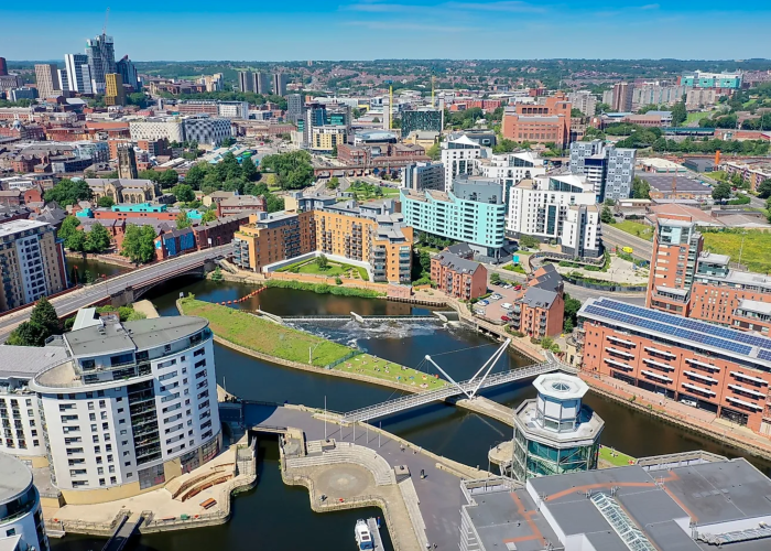 Vue aérienne d'un paysage urbain moderne avec une rivière, des ponts piétonniers et des bâtiments contemporains entourés de verdure sous un ciel bleu clair. La ville s'étend au loin avec d'autres bâtiments et collines visibles.