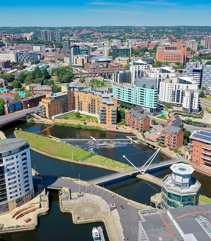 Vue aérienne d'un paysage urbain moderne avec une rivière, des ponts piétonniers et des bâtiments contemporains entourés de verdure sous un ciel bleu clair. La ville s'étend au loin avec d'autres bâtiments et collines visibles.