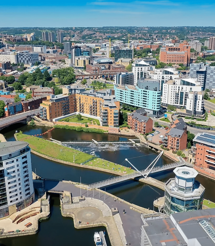 Vue aérienne d'un paysage urbain moderne avec une rivière, des ponts piétonniers et des bâtiments contemporains entourés de verdure sous un ciel bleu clair. La ville s'étend au loin avec d'autres bâtiments et collines visibles.