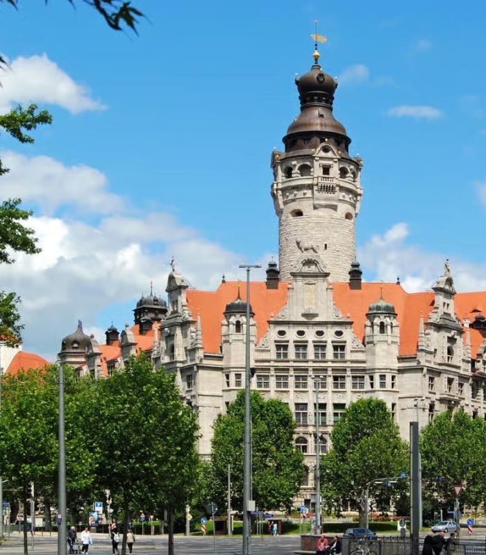 Un bâtiment historique en pierre avec une haute tour centrale et un toit en tuiles rouges se dresse derrière des arbres verts sous un ciel bleu parsemé de nuages.
