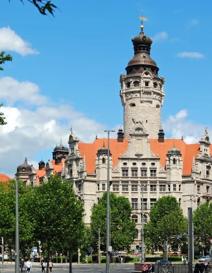 Un bâtiment historique en pierre avec une haute tour centrale et un toit en tuiles rouges se dresse derrière des arbres verts sous un ciel bleu parsemé de nuages.