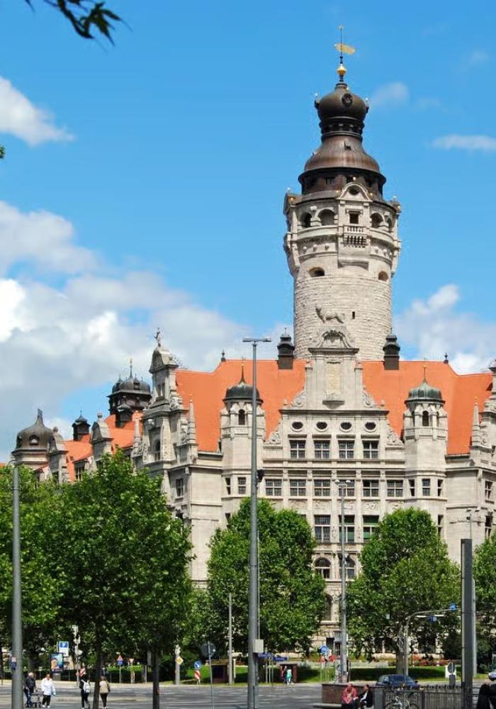 A historic stone building with a tall central tower and red-tiled roof stands behind green trees under a blue sky dotted with clouds.
