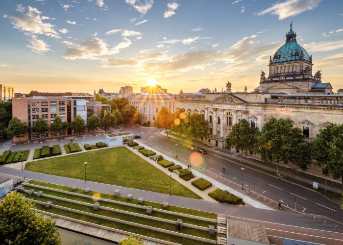 Un paysage urbain au coucher du soleil présente un bâtiment historique en forme de dôme à côté d'un grand parc verdoyant et de structures modernes. La lumière du soleil traverse les nuages et jette une lueur chaude sur les arbres et les rues vides.