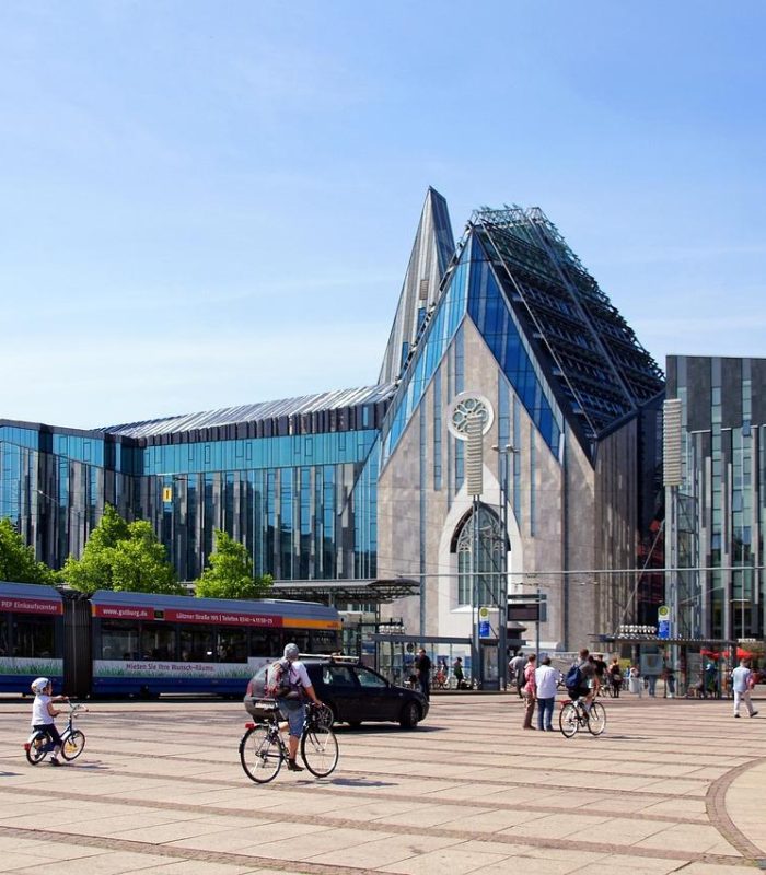 Modern glass buildings with unique angular architecture on a city square, people on foot and on bikes, trams and buses in the street, under a clear blue sky.