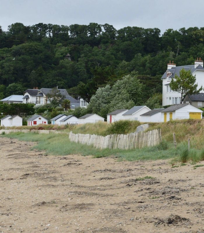 A sandy beach bordered by grassy dunes and a wooden fence, with small white beach huts and houses nestling among the trees in the background, under a cloudy sky.