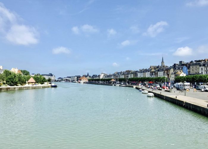 A wide river lined with boats flows through a town with historic buildings and a church spire. Trees and a promenade line the right bank under a bright, partly cloudy sky.