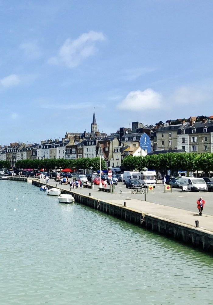Un front de mer avec des bateaux amarrés le long d'une jetée, des gens marchant sur le trottoir, et des rangées de bâtiments colorés avec des magasins et des restaurants sous un ciel bleu. Un clocher d'église se dresse à l'arrière-plan.