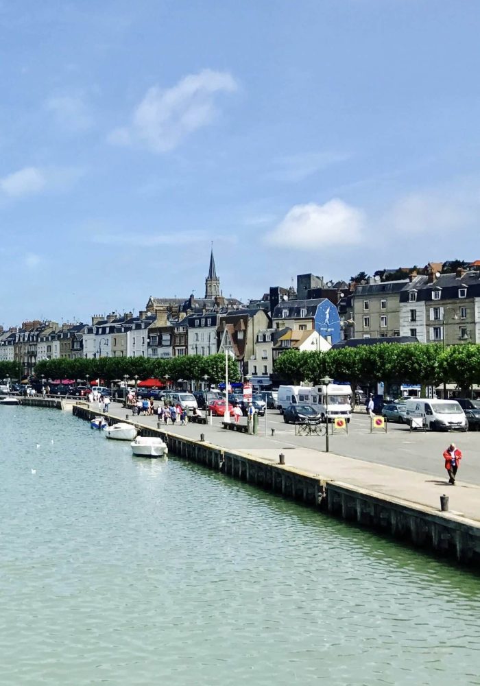 A seafront with boats moored along a pier, people walking along the pavement, and rows of colourful buildings with shops and restaurants under a blue sky. A church tower stands in the background.