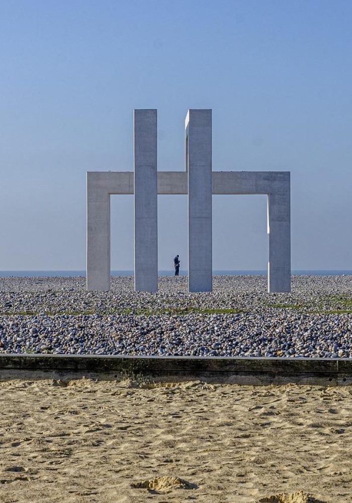 Une grande sculpture géométrique en béton se dresse sur une plage rocheuse sous un ciel bleu clair, avec une seule personne visible au loin à travers la structure. Le sable est au premier plan.