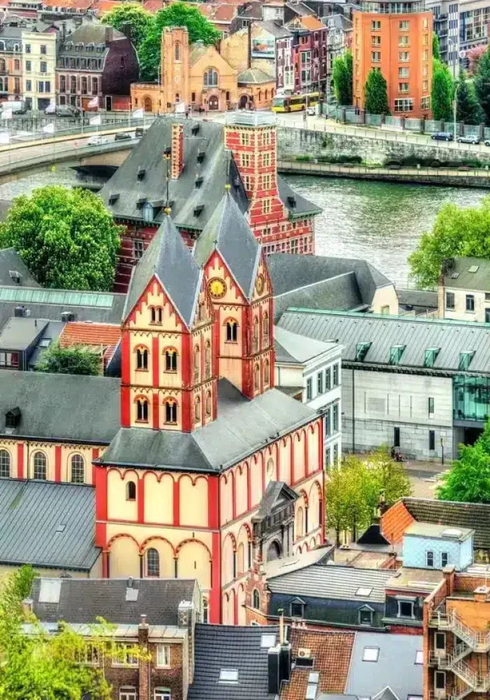 Colourful historic buildings surround a large church with red and white accents in a European town on the banks of a river, with a bridge and green trees in the background.