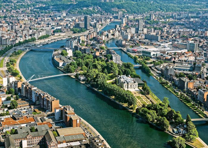Vue aérienne d'une ville avec une rivière qui se divise en deux branches, des îles bordées d'arbres, des ponts et un mélange de bâtiments modernes et historiques. Des parcs et des espaces verts sont visibles le long des berges.