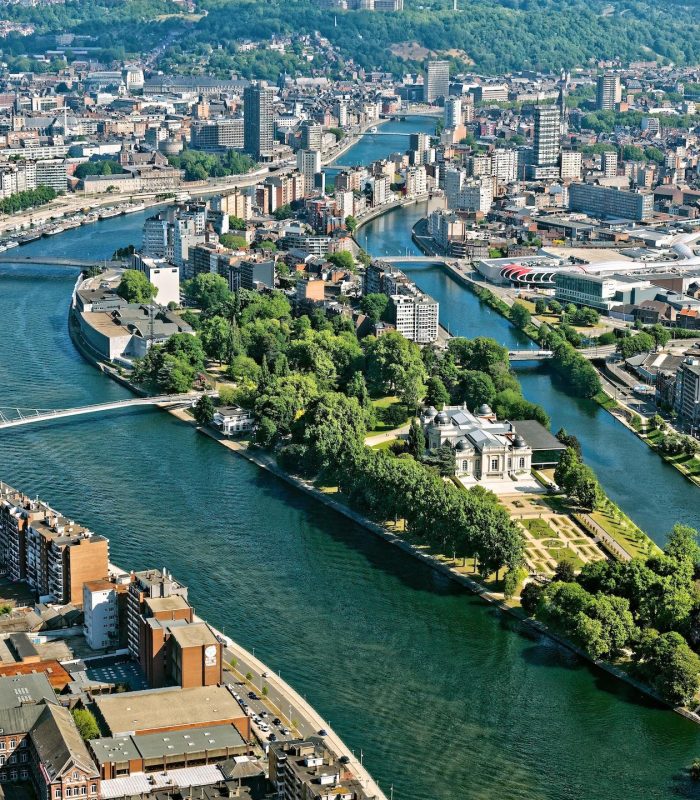 Vue aérienne d'une ville avec une rivière qui se divise en deux branches, des îles bordées d'arbres, des ponts et un mélange de bâtiments modernes et historiques. Des parcs et des espaces verts sont visibles le long des berges.