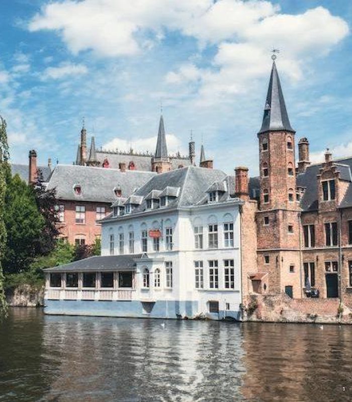 Un canal pittoresque à Bruges, en Belgique, bordé de bâtiments historiques en brique et d'une église gothique, avec un arbre feuillu sur la gauche et des reflets dans l'eau sous un ciel partiellement nuageux.