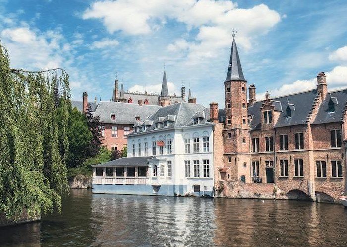 A picturesque canal in Bruges, Belgium, lined with historic brick buildings and a Gothic church, with a leafy tree on the left and reflections in the water under a partly cloudy sky.