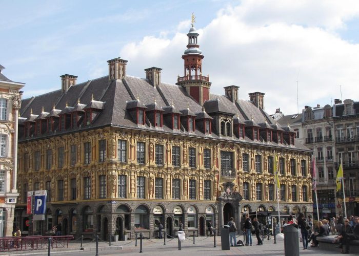 A large historic building adorned with yellow stonework, arched windows and a central tower stands in a busy square where people stroll and others sit by a fountain. The sky is partly cloudy.