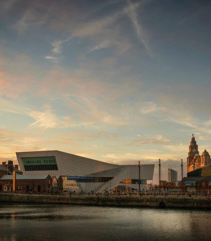Liverpool's waterfront at sunset, with modern and historic buildings, including the Museum of Liverpool and the Royal Liver Building, reflected in the calm waters of the River Mersey under a colourful sky.