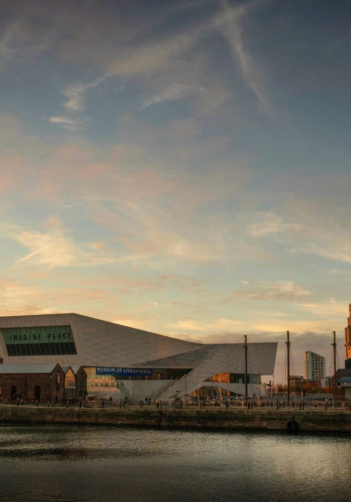 Liverpool's waterfront at sunset, with modern and historic buildings, including the Museum of Liverpool and the Royal Liver Building, reflected in the calm waters of the River Mersey under a colourful sky.
