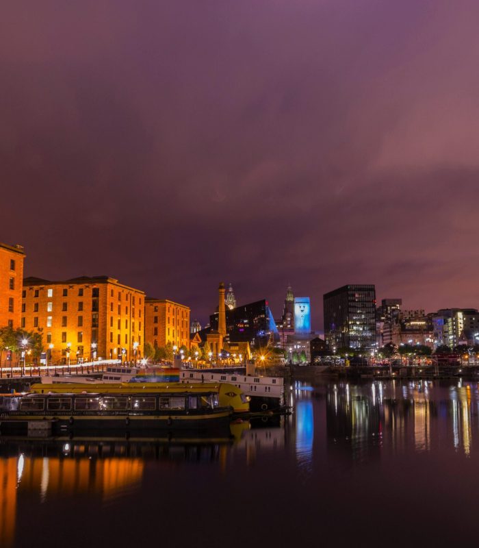 Vue nocturne du front de mer d'une ville avec, à gauche, des bâtiments historiques en briques brillamment éclairés, à droite, des gratte-ciel modernes et des bateaux amarrés dans une eau calme reflétant les lumières colorées.