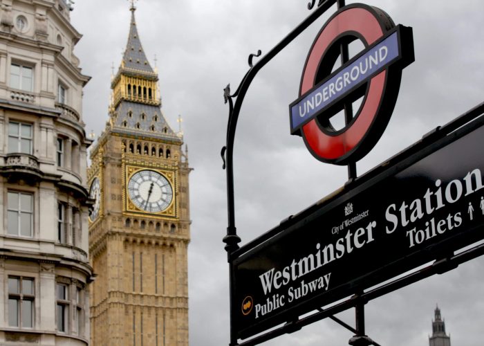 Un panneau du métro londonien indiquant la station Westminster se trouve au premier plan, tandis que la tour de l'horloge Big Ben et une partie d'un bâtiment historique sont visibles dans un ciel nuageux à l'arrière-plan.