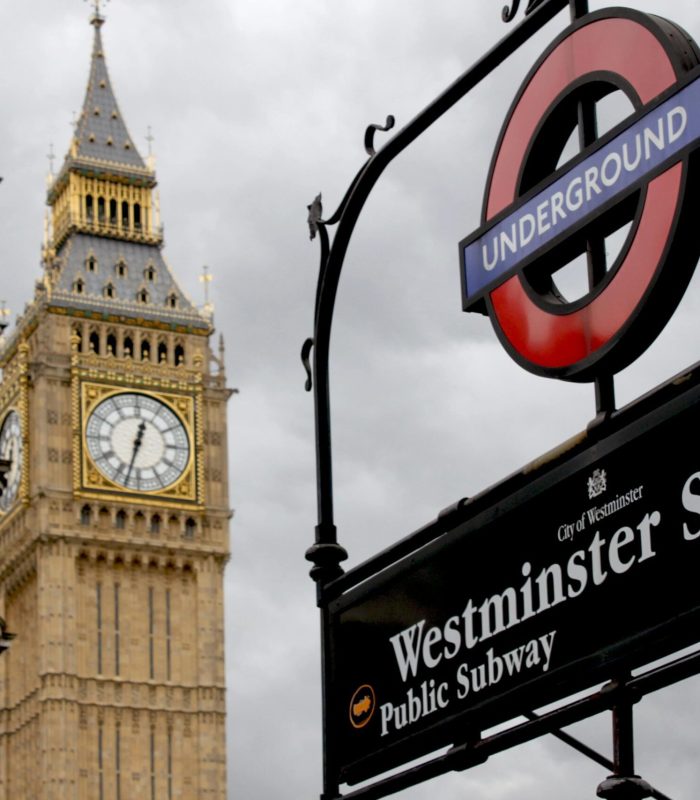 Un panneau du métro londonien indiquant la station Westminster se trouve au premier plan, tandis que la tour de l'horloge Big Ben et une partie d'un bâtiment historique sont visibles dans un ciel nuageux à l'arrière-plan.