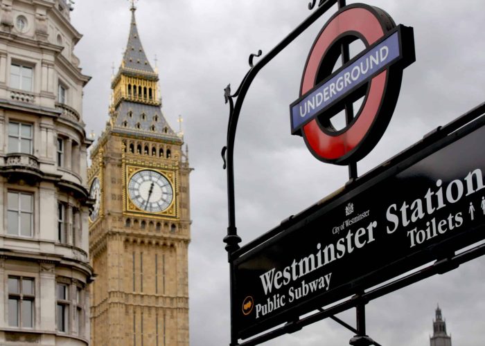 A London Underground sign pointing to Westminster station is in the foreground, while the Big Ben clock tower and part of a historic building can be seen against a cloudy sky in the background.