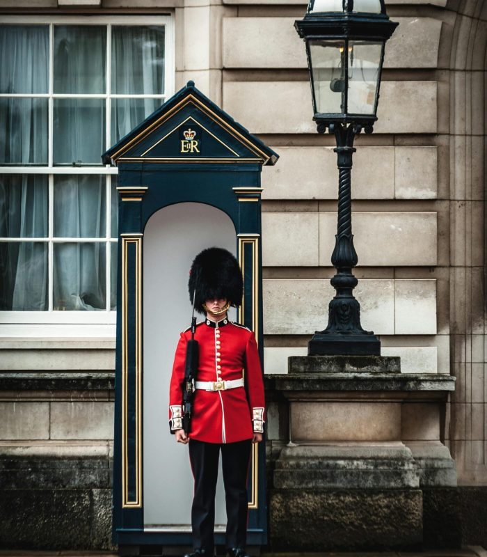 Un garde royal britannique en uniforme rouge et bonnet de peau d'ours se tient au garde-à-vous à l'extérieur d'une guérite, à côté d'un mur de pierre et d'une lampe ancienne.
