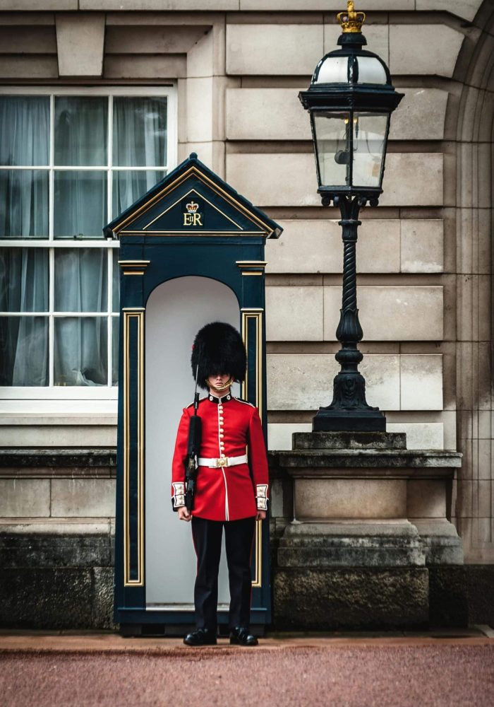 A British Royal Guardsman in red uniform and bearskin cap stands at attention outside a sentry box, next to a stone wall and an antique lamp.
