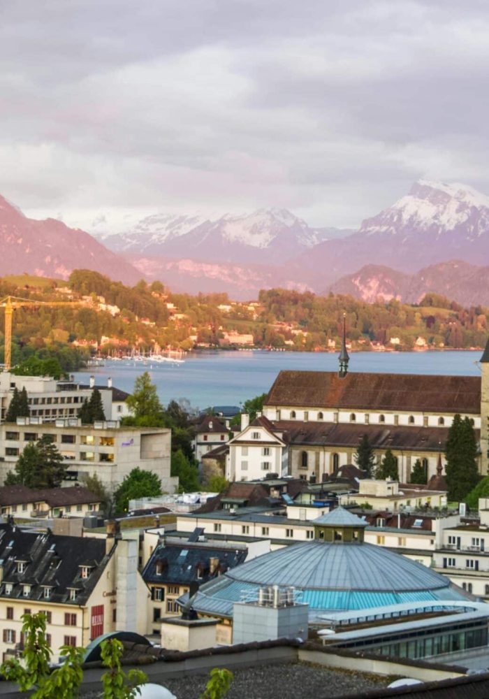 Vue panoramique d'une ville européenne avec des bâtiments historiques, une église avec deux grandes flèches, un lac et des montagnes enneigées en arrière-plan sous un ciel nuageux.