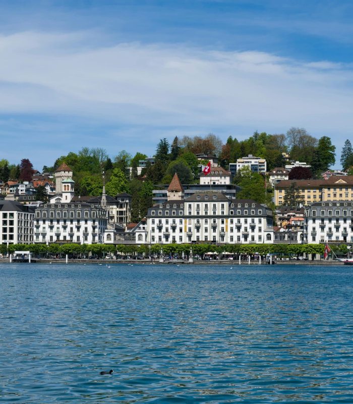 Vue panoramique de bâtiments historiques et d'hôtels le long du front de mer à Lucerne, en Suisse, avec une colline couverte d'arbres en arrière-plan sous un ciel bleu partiellement nuageux.