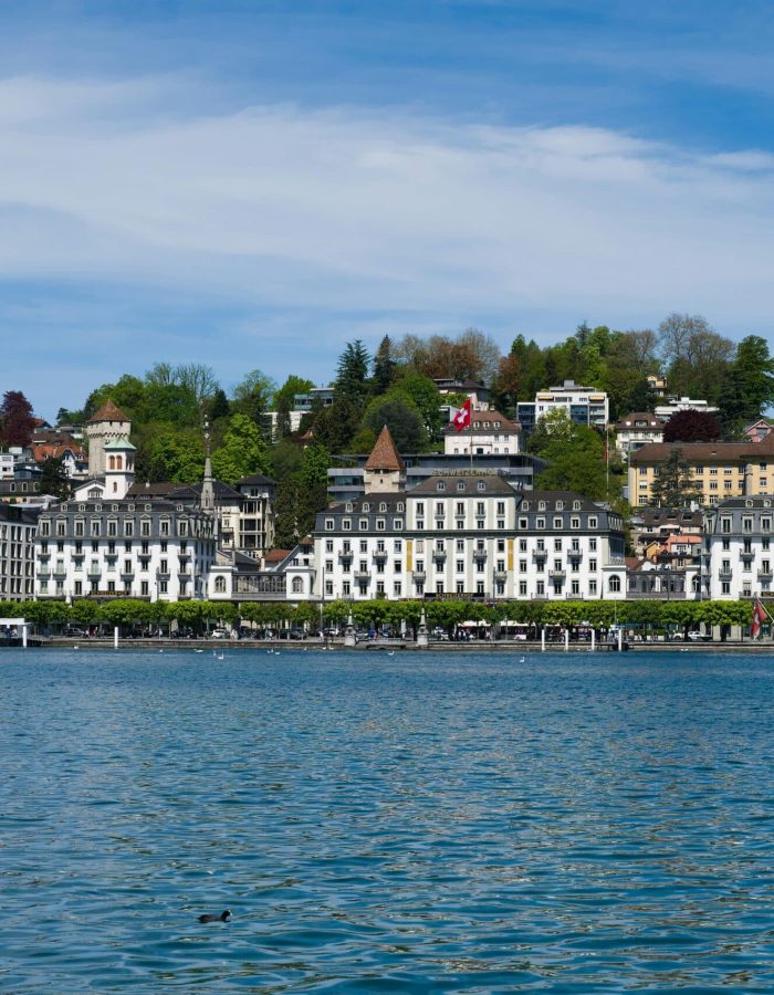 Vue panoramique de bâtiments historiques et d'hôtels le long du front de mer à Lucerne, en Suisse, avec une colline couverte d'arbres en arrière-plan sous un ciel bleu partiellement nuageux.