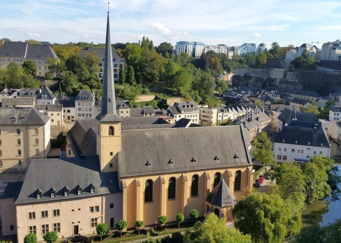 Vue aérienne d'une ville européenne historique avec une grande église dotée d'une haute flèche, entourée de vieux bâtiments, d'arbres et d'une rivière sous un ciel bleu clair.
