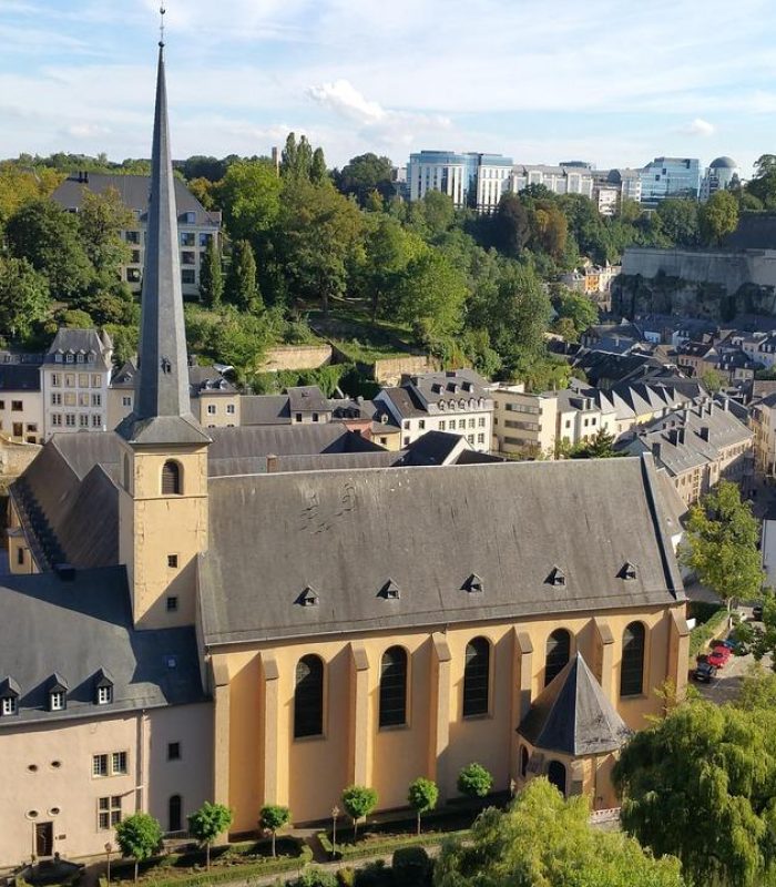 Vue aérienne d'une ville européenne historique avec une grande église dotée d'une haute flèche, entourée de vieux bâtiments, d'arbres et d'une rivière sous un ciel bleu clair.