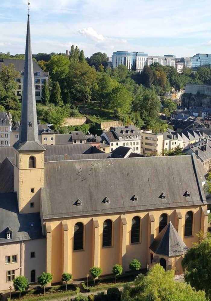 Vue aérienne d'une ville européenne historique avec une grande église dotée d'une haute flèche, entourée de vieux bâtiments, d'arbres et d'une rivière sous un ciel bleu clair.