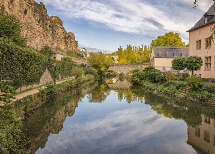 Une rivière calme bordée de murs de pierre, de verdure luxuriante et de vieux bâtiments, reflétant le ciel et le paysage environnant ; des falaises rocheuses s'élèvent sur la gauche et un pont de pierre enjambe la rivière à l'arrière-plan.