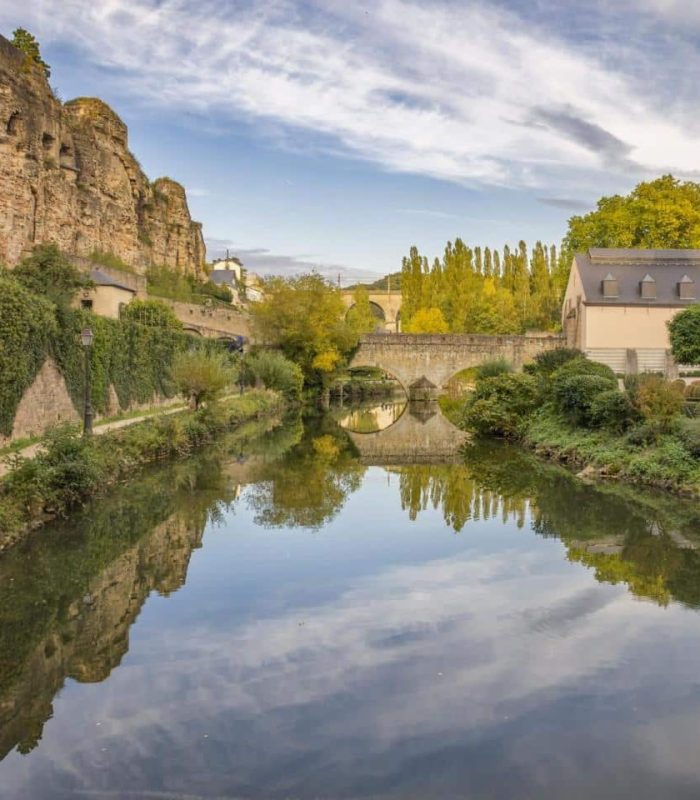 Une rivière calme bordée de murs de pierre, de verdure luxuriante et de vieux bâtiments, reflétant le ciel et le paysage environnant ; des falaises rocheuses s'élèvent sur la gauche et un pont de pierre enjambe la rivière à l'arrière-plan.
