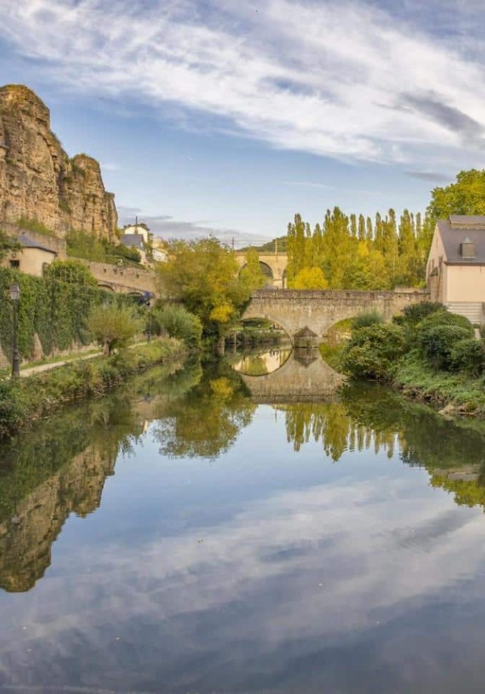 Une rivière calme bordée de murs de pierre, de verdure luxuriante et de vieux bâtiments, reflétant le ciel et le paysage environnant ; des falaises rocheuses s'élèvent sur la gauche et un pont de pierre enjambe la rivière à l'arrière-plan.