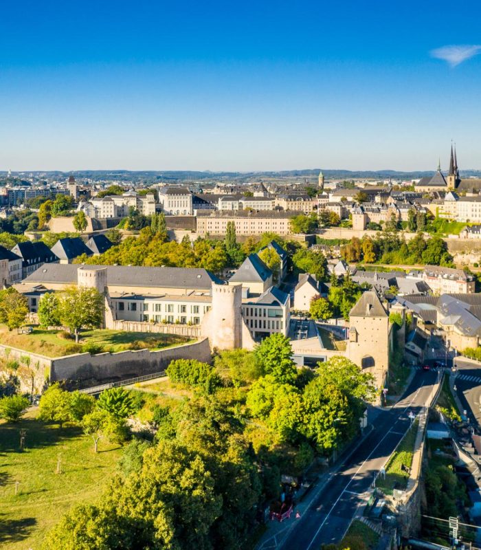 Vue aérienne de la ville de Luxembourg montrant des fortifications historiques, des bâtiments en pierre, des arbres verdoyants et un ciel bleu clair par une journée ensoleillée.