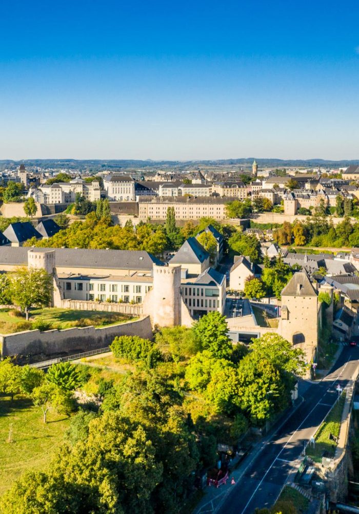Vue aérienne de la ville de Luxembourg montrant des fortifications historiques, des bâtiments en pierre, des arbres verdoyants et un ciel bleu clair par une journée ensoleillée.